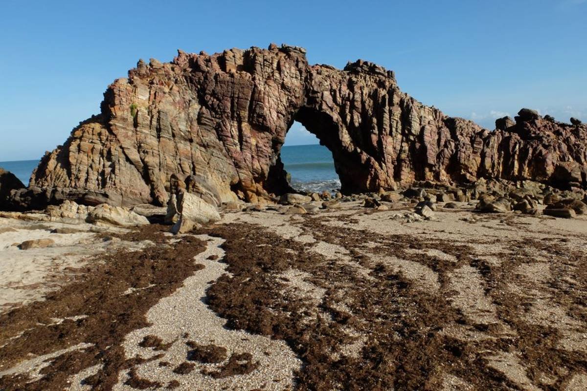 Foto colorida de rocha gigante com furo ao meio em meio a uma praia - Metrópoles