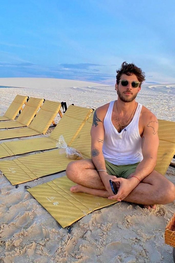 Foto colorida de homem branco e de óculos sentado em espreguiçadeira colocada em areia - Metrópoles