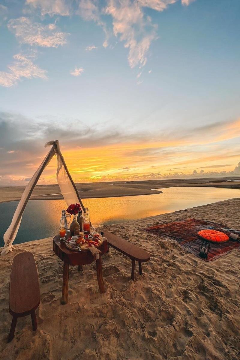 Foto colorida de pôr do sol, dunas, rio e cabana montada com mesa e cadeira - Metrópoles