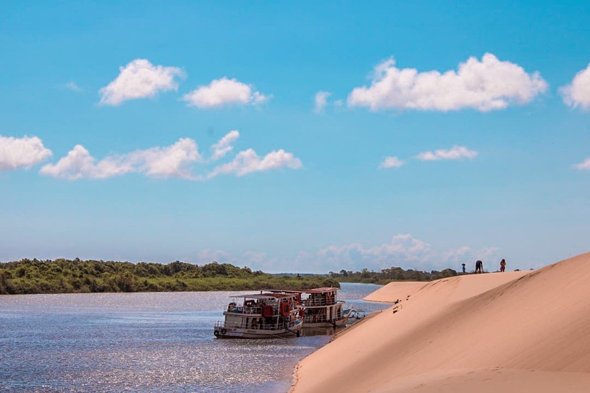 Foto colorida de barco em rio com dunas. O tempo está ensolarado - Metrópoles