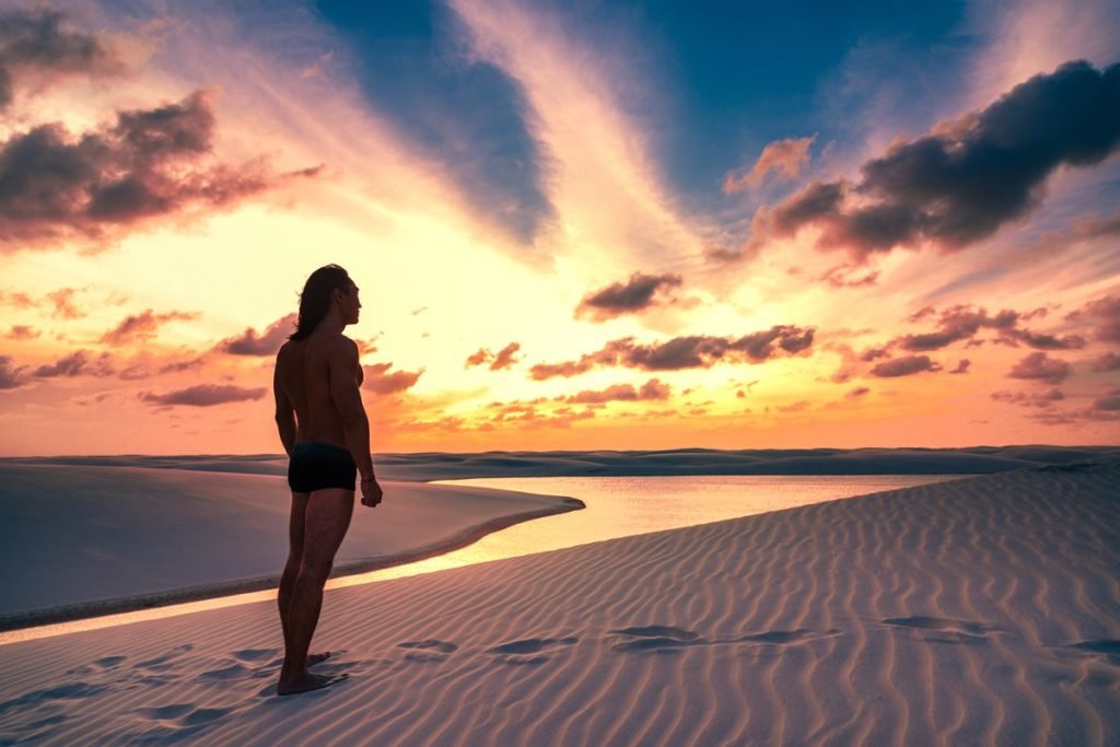 Foto colorida de homem em duna observando o pôr do sol - Metrópoles