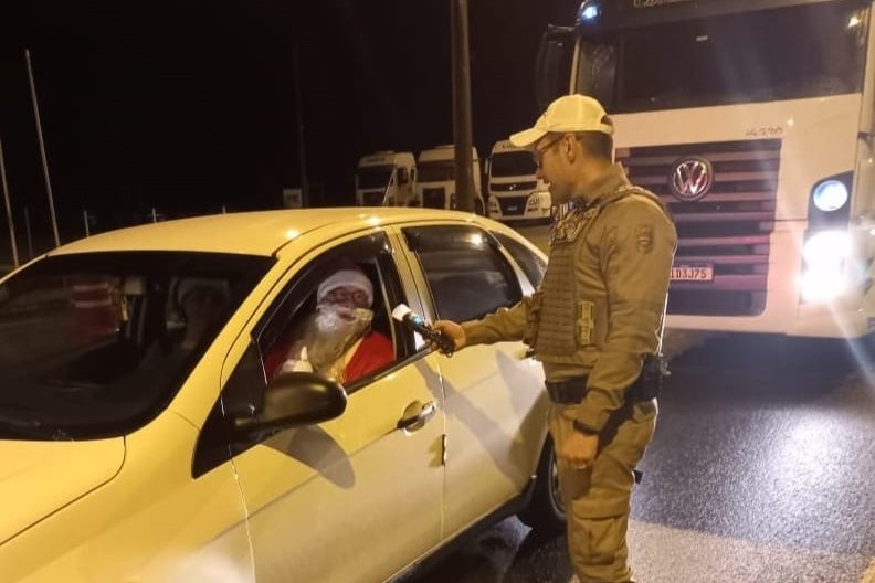 Imagem colorida: homem vestido de Papai Noel faz teste do bafômetro dentro de carro, ao lado de policial