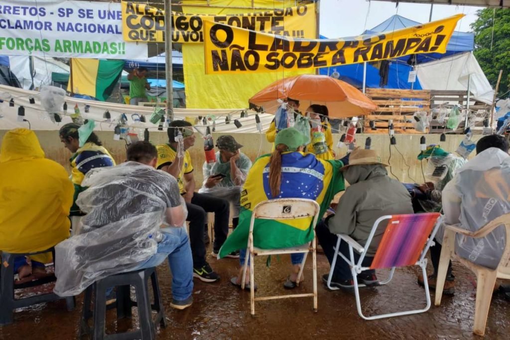 Registros de barracas arrasadas pela chuva no acampamento de militantes bolsonaristas em frente ao QG do Exército de Brasília, com muita lama em volta. No destaque, manifestantes aparecem juntos debaixo de lona com capas de chuva e faixas - Metrópoles