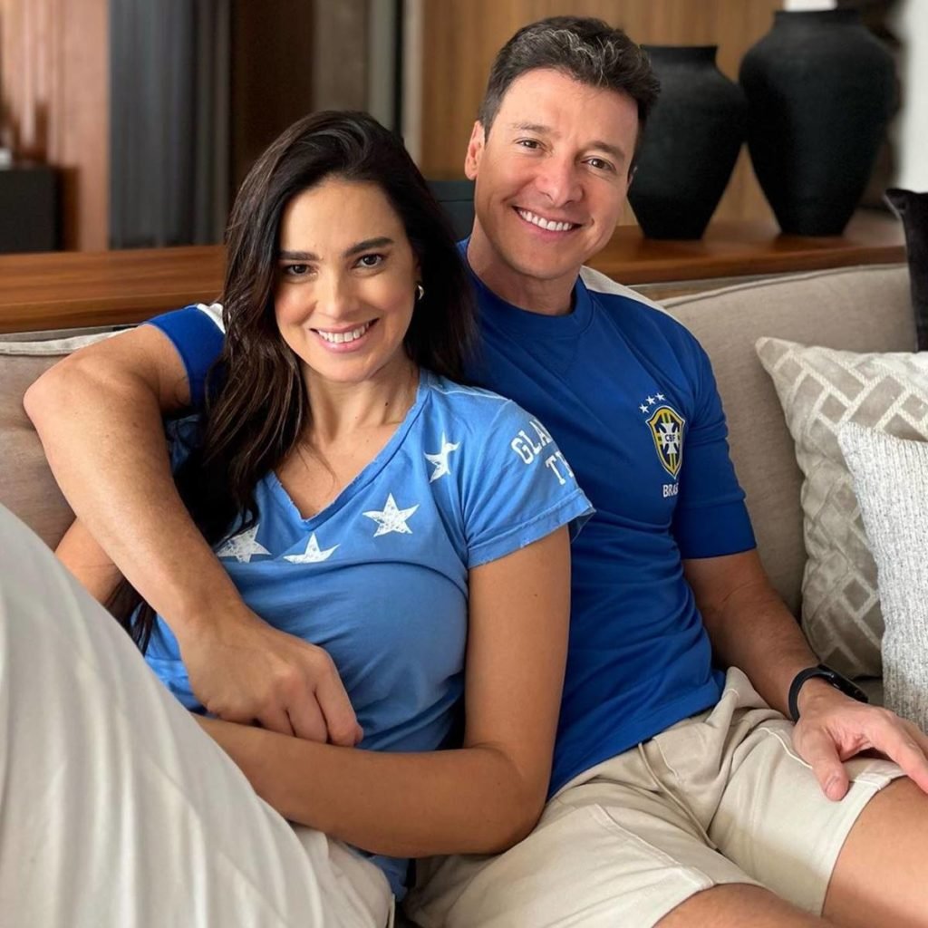 Rodrigo Faro sentado no sofá de blusa do brasil e bermuda branca abraçado na esposa Vera Viel, de camiseta azul com estrelas e calça branca - metrópoles