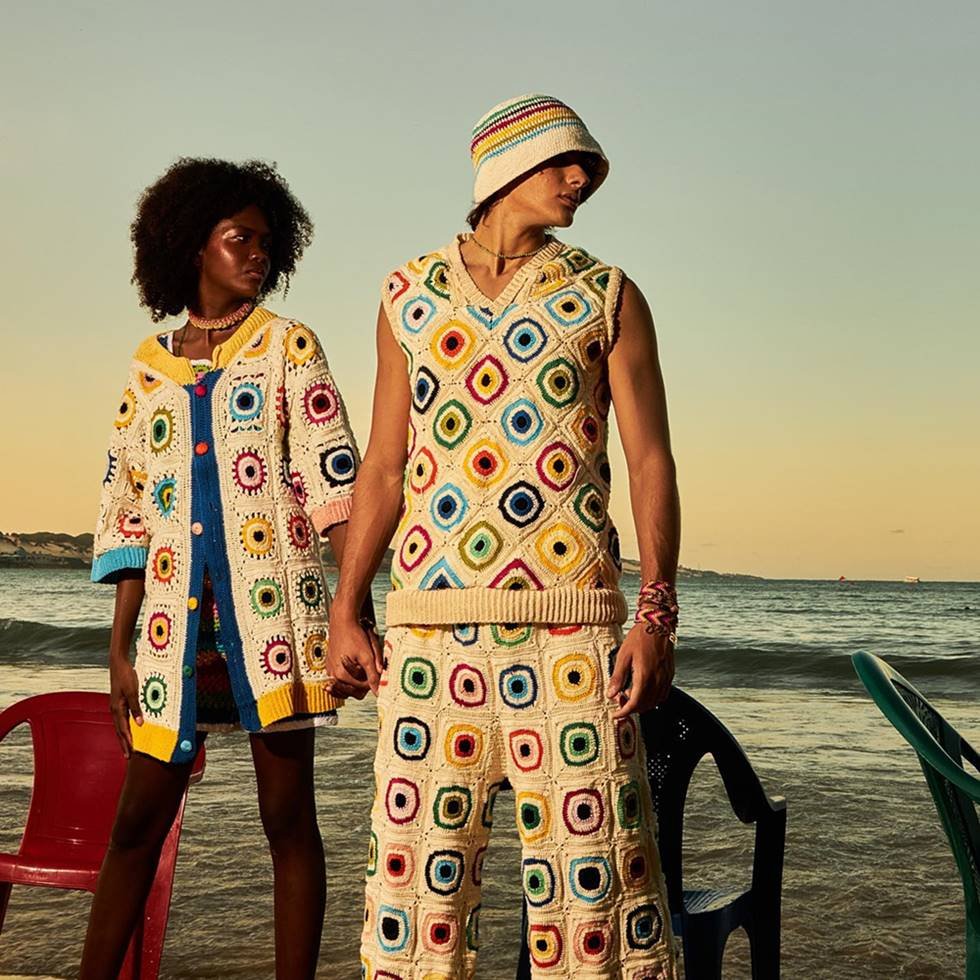 Uma mulher negra e um homem branco, ambos jovens e modelos, posam em uma praia para uma campanha de fotos da marca Depedro. Ela usa um vestido e ele usa um conjunto de blusa e calça. Todas as peças são de crochê branco com detalhes coloridos. - Metrópoles