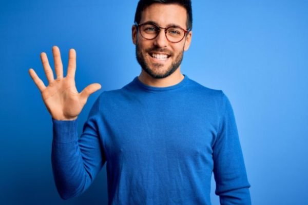 Homem com blusa azul fazendo o numeral 5 com a mão e sorrindo