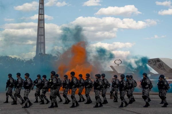 Agentes da Força Nacional marcham na Praça dos Três Poderes uniformizados com explosões de pó colorido ao fundo - Metrópoles
