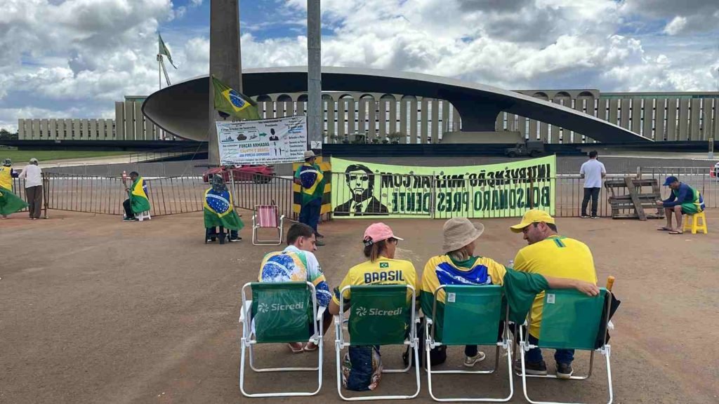 Acampamento bolsonarista no QG do Exército, em Brasília, com manifestantes sentados diante de imenso monumento em frente de prédio - Metrópoles