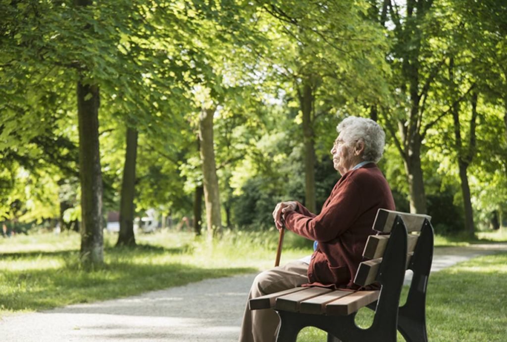 Imagem colorida: idosa solitória sentada em banco de parque com uma bengala - Metrópoles