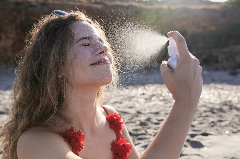 Mulher loira aplicando spray em rosto com praia ao fundo - Metrópoles