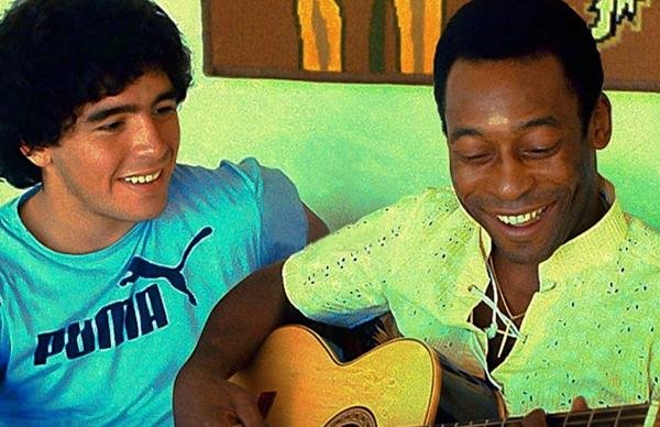 Os jogadores Maradona (Argentina) e Pelé (Brasil) sorriem e cantam enquanto Pelé toca um violão - Metrópoles