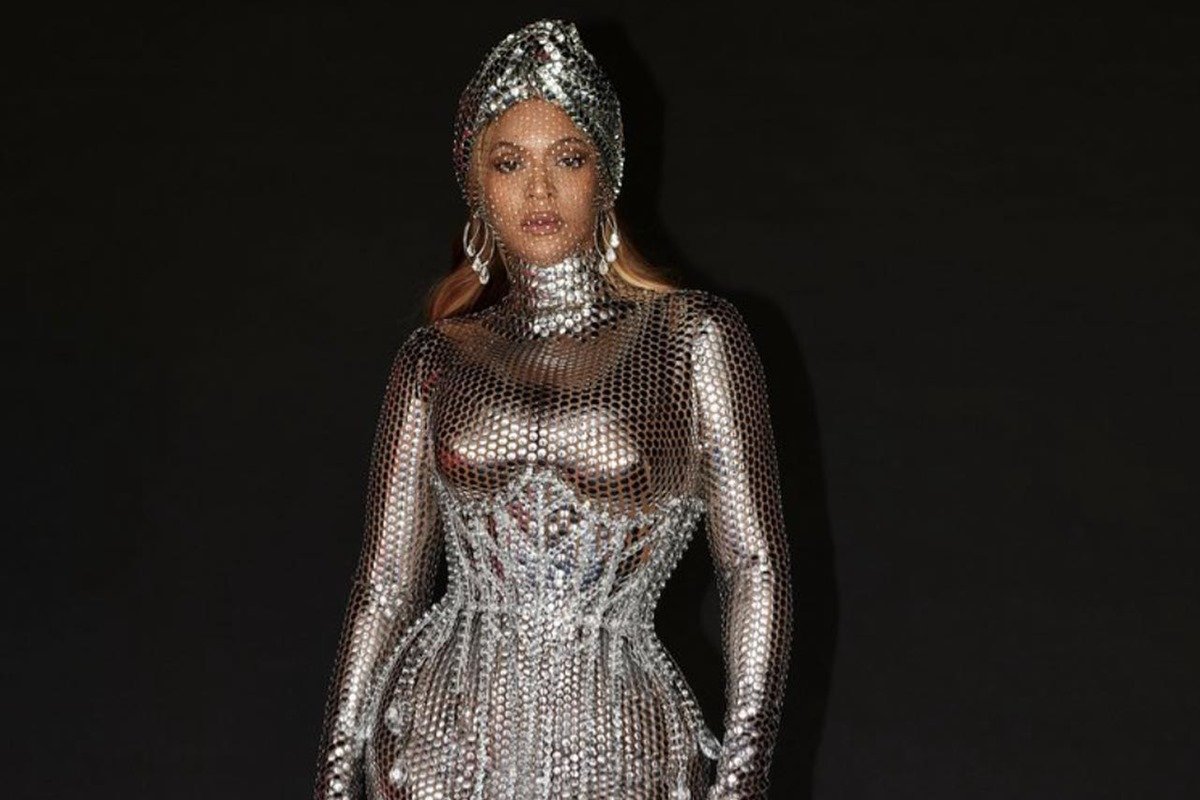 A cantora Beyoncé, uma mulher negra e de meia idade, com o cabelo liso e loiro, posa para foto em um fundo preto. Ela usa um vestido prata e um turbante na cabeça, também prata. As peças são da marca de moda Burberry. - Metrópoles