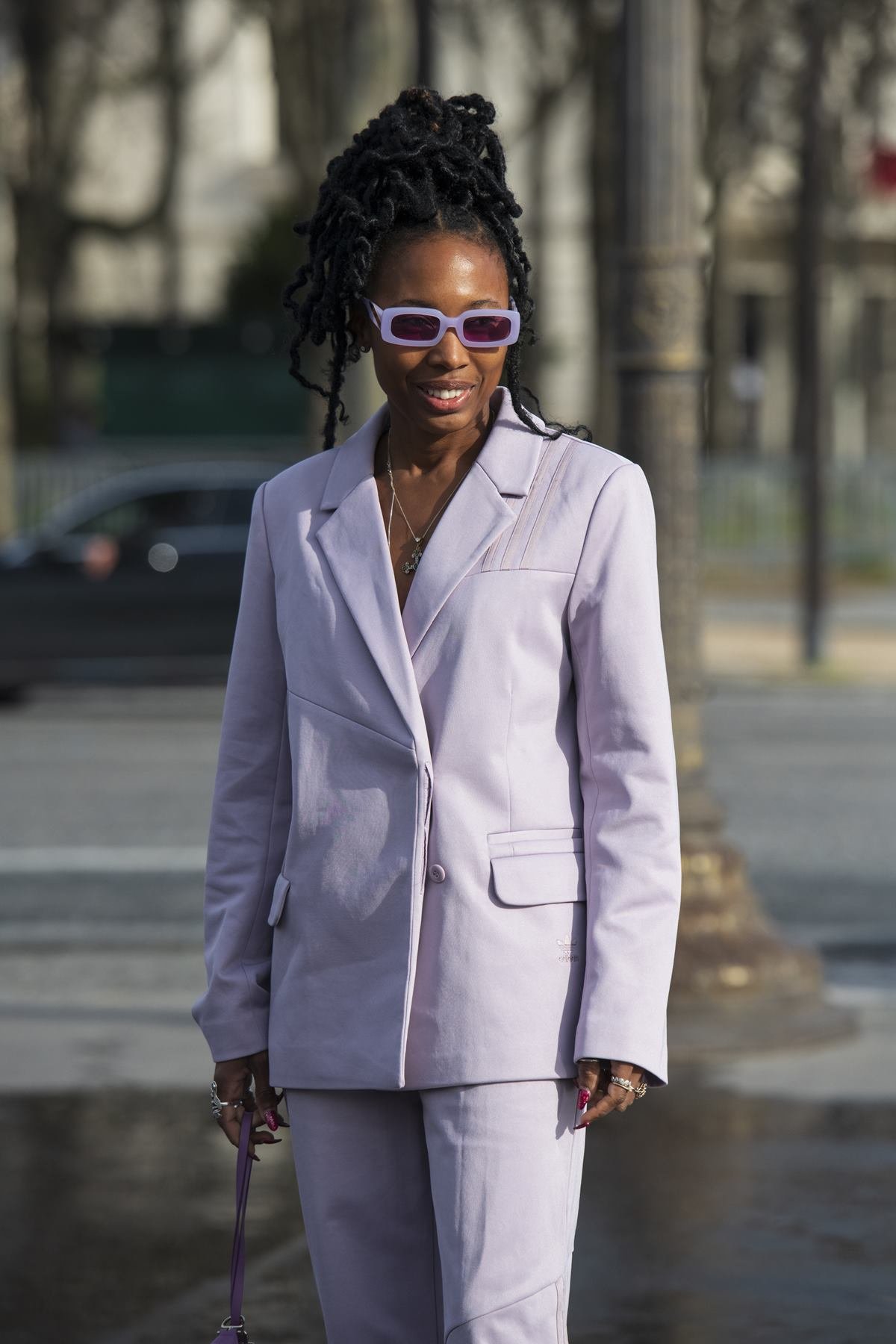 A stylist Zerina Akers, uma mulher negra e jovem, de cabelo trançado amarrado, posa para foto nas ruas de Paris durante a semana de moda. Ela usa um conjunto de blazer e calça, ambos na cor lilás, e um óculos de sol com armação roxa. - Metrópoles