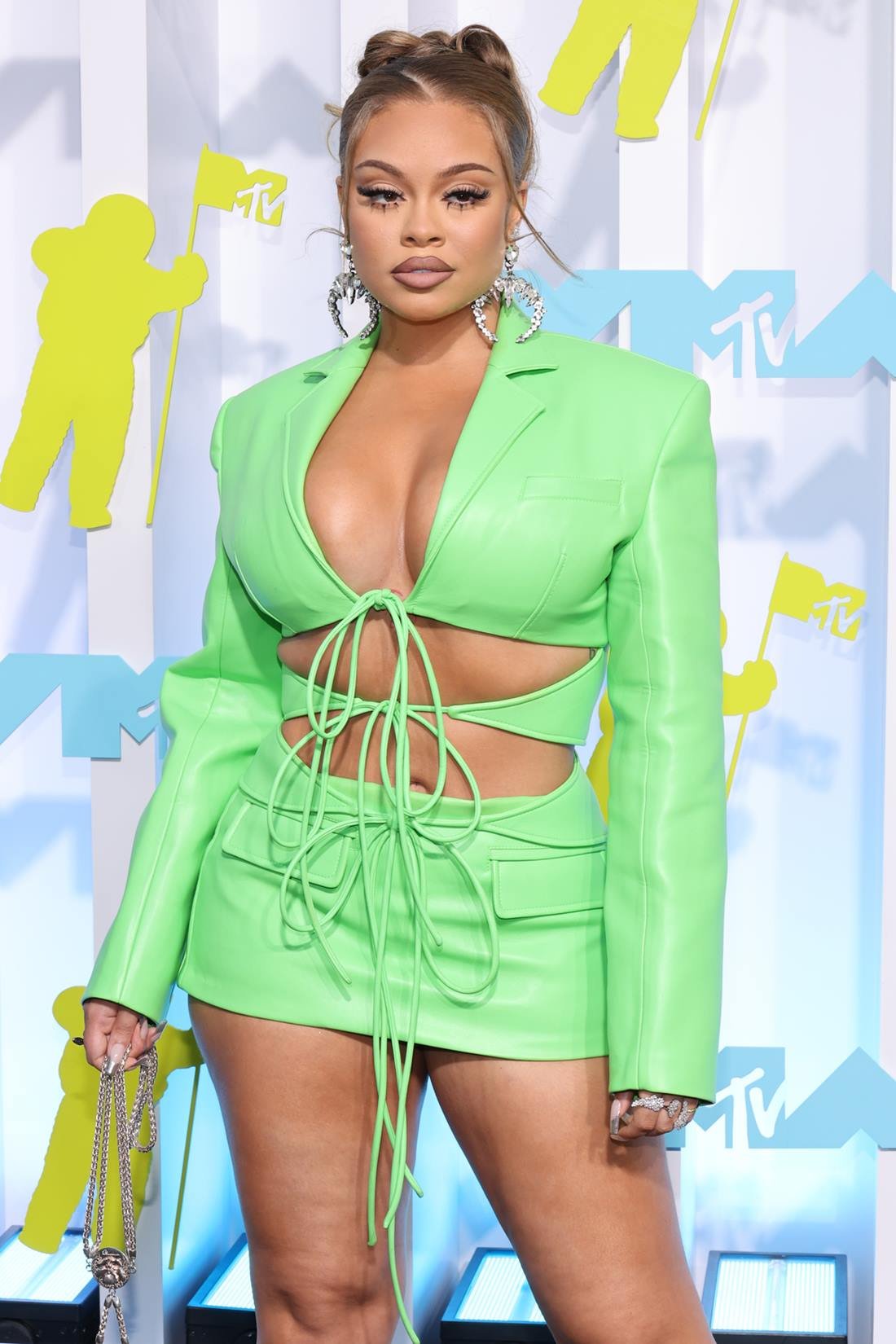 A cantora Big Latto, uma mulher branca e jovem, de cabelo liso loiro, posa para foto na premiação MTV Awards. Ela usa um vestido estilo blazer com recortes e amarrações na cor verde e um brinco de argola dourada. - Metrópoles
