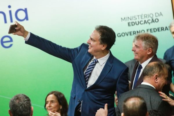 Camilo Santana toma posse como ministro da Educação em cerimônia no Ministério, cercado de autoridades e políticos. Ele tira selfie com outro homem e sorri na imagem - Metrópoles