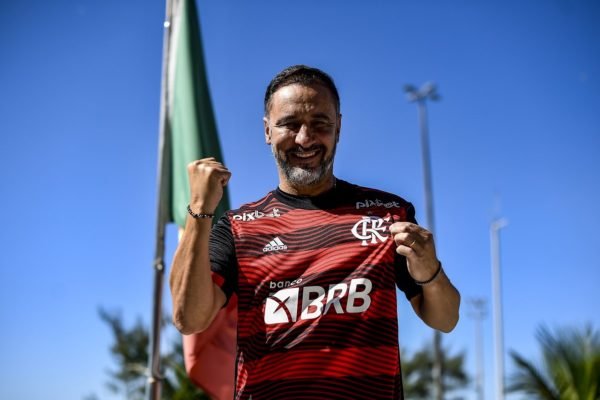 Novo técnico do Flamengo, Vítor Pereira, sorri, ergue o punho em riste e segura logo do time na camisa em foto - Metrópoles