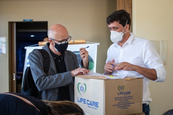 Dois homens de máscara assinam papéis que estão em cima de uma caixa de papelão que contém máscaras