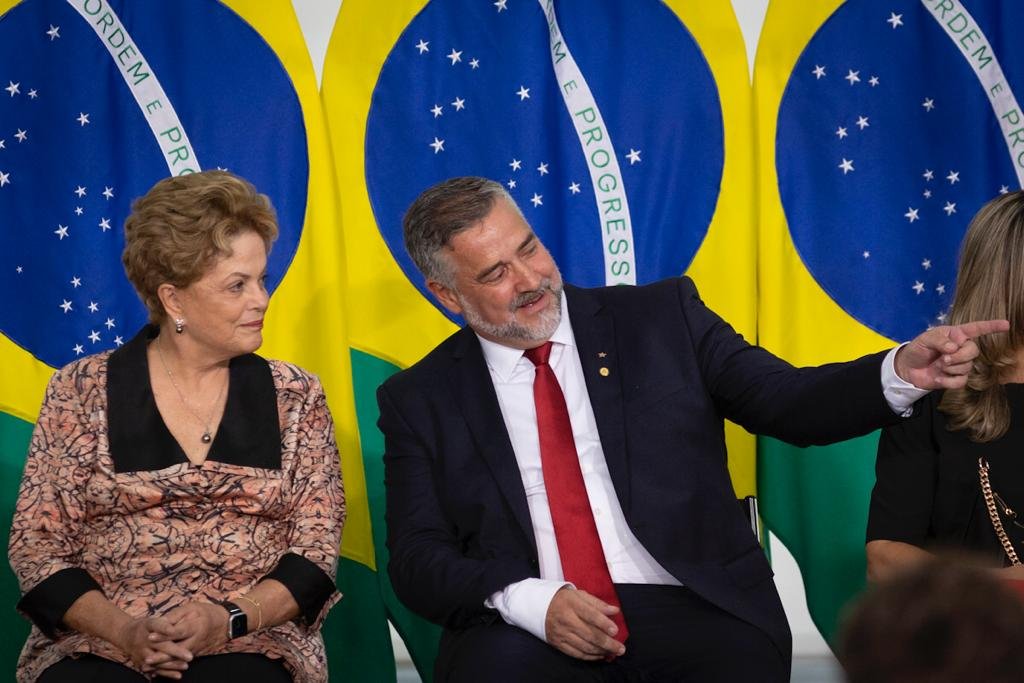 Paulo Pimenta toma posse como ministro de Estado Chefe da Secretaria de Comunicação da Presidência da República em cerimônia no palácio do Planalto. Ele conversa, sentado ao lado da ex-presidente Dilma Rousseff, com bandeiras do Brasil atrás - Metrópoles