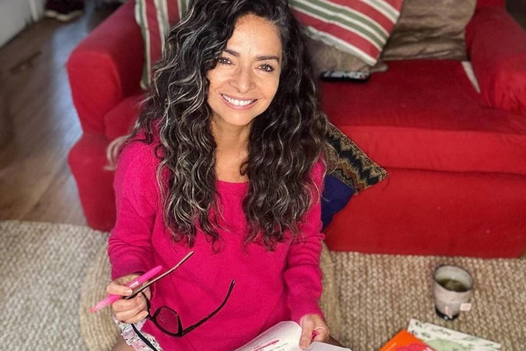 mulher de cabelo cacheado e blusa de frio rosa sentada no tapete enquanto segura óculos de grau - metrópoles