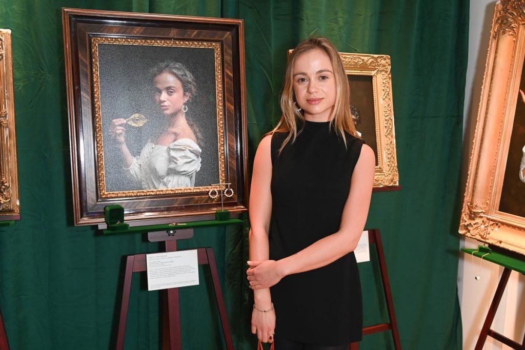Foto colorida de mulher branca e loira com vestido preto em frente a um quadro que a retrata - Metrópoles
