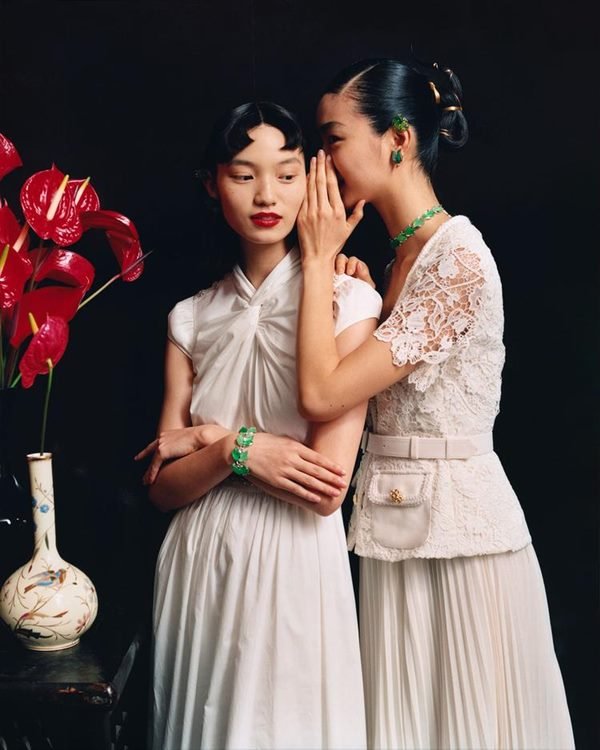 Duas mulheres asiáticas em editorial de moda. Ambas estão usando roupas brancas e uma está falando no ouvido da outra - Metrópoles