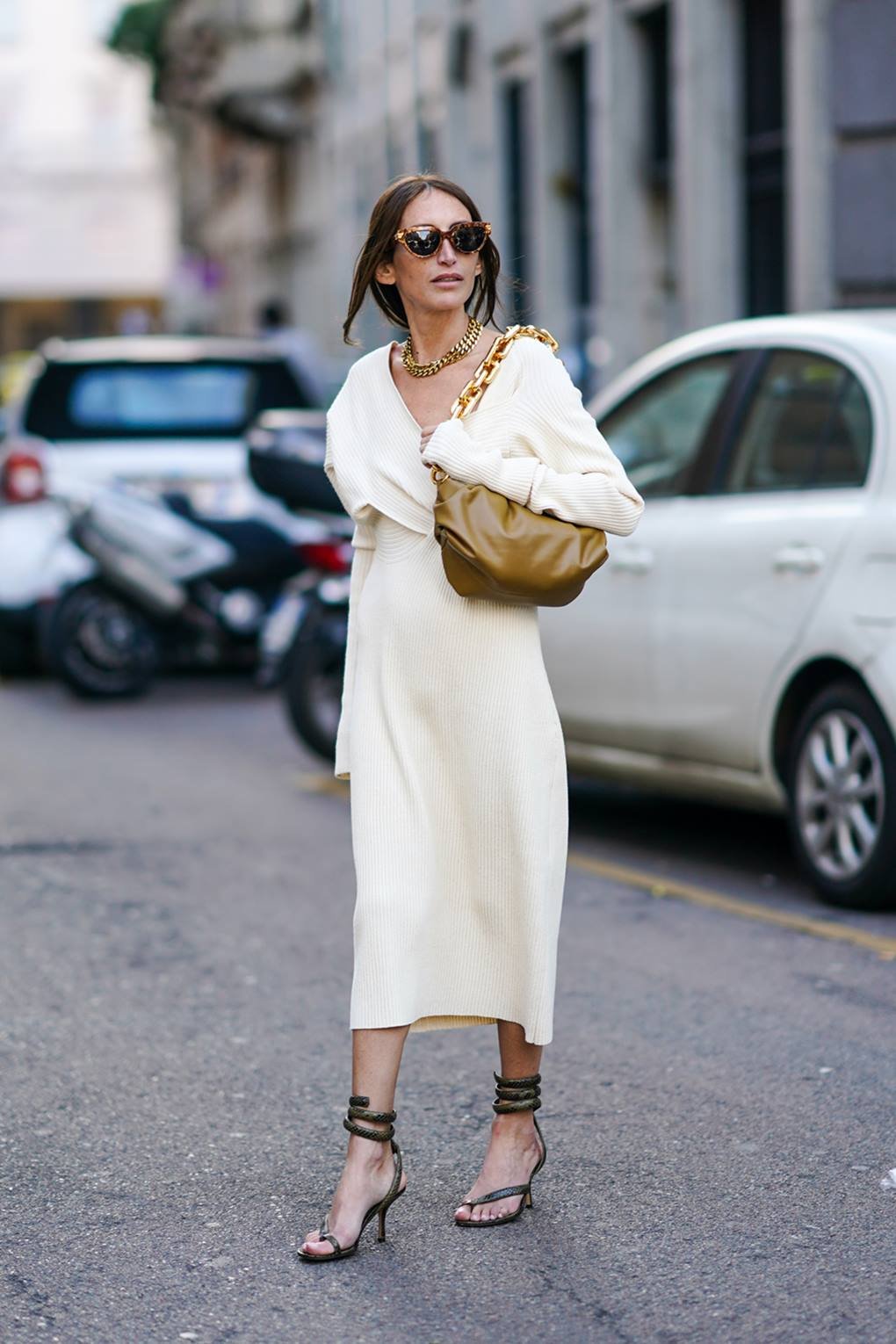 Mulher branca e jovem, de cabelo castanho liso, andando pelas ruas de Paris, na França. Ela usa um vestido branco midi, com mangas longas, uma bolsa marrom de couro, óculos escuros e uma sandália de salto preta. - Metrópoles