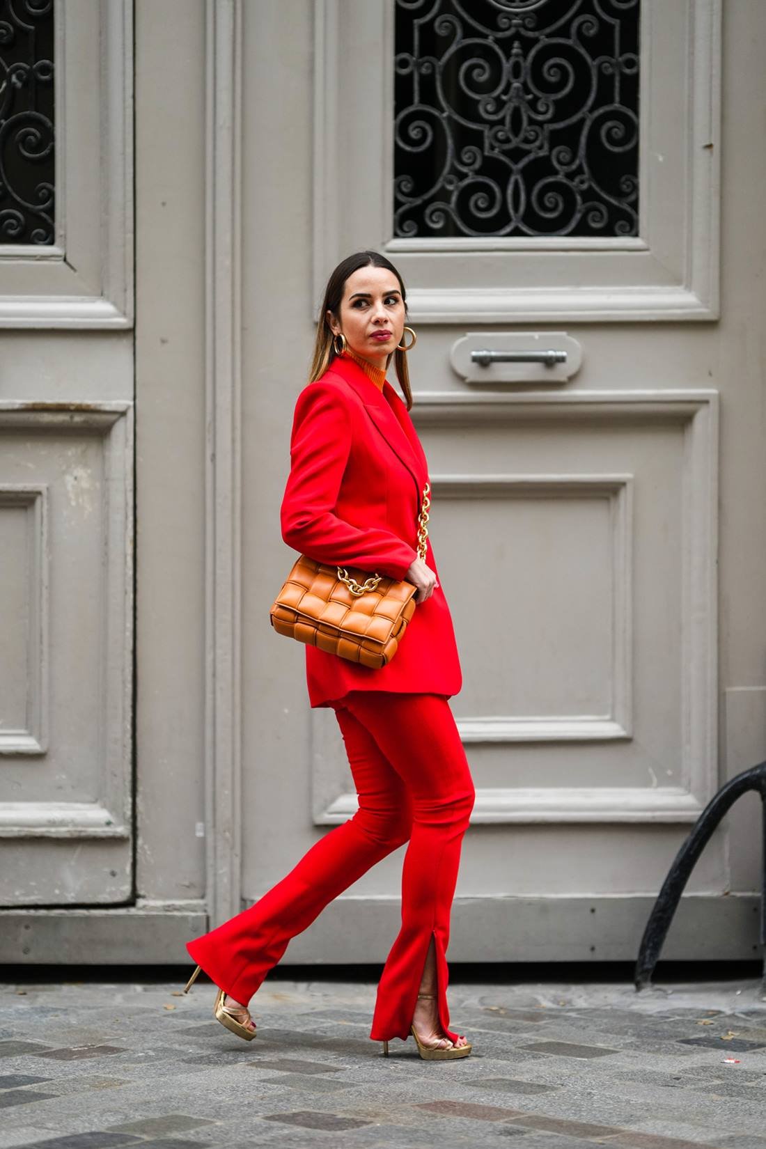 Mulher branca e jovem, de cabelo liso e loiro, andando pelas ruas de Paris, na França. Ela usa um conjunto de blazer e calça social, ambos vermelho, uma sandália de salto e uma bolsa de couro marrom transpassada. - Metrópoles