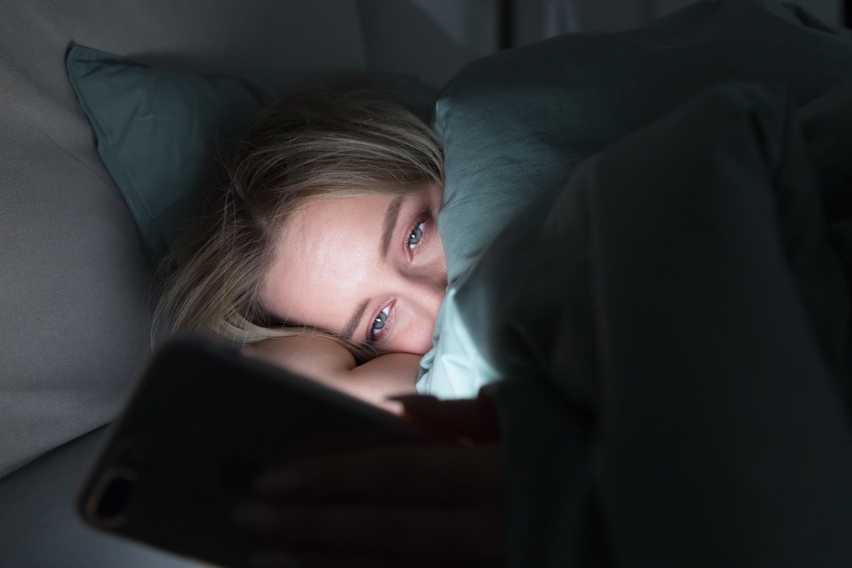 Foto colorida de mulher branca e loira mexendo no celular deitada em uma cama - Metr&oacute;poles