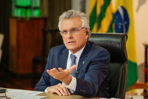 O governador de Goiás, Ronaldo Caiado, durante entrevista virtual, sentado diante de mesa em sua sala. Ele usa ponto eletrônico no ouvido com bandeiras atrás - Metrópoles