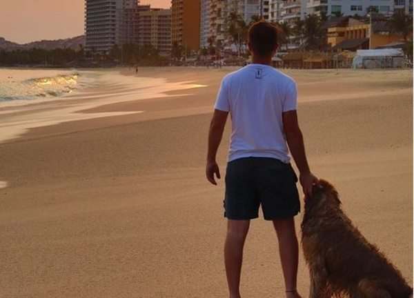 Foto colorida de um homem de camisa branca e bermuda em uma praia ao lado de um cachorro