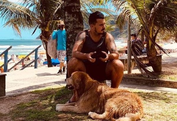 Foto colorida de um homem sentado em baixo de um coqueiro na praia ao lado de um cachorro