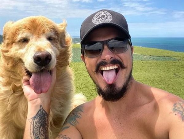 Foto colorida de um homem de óculos escuros e boné ao lado de um cachorro