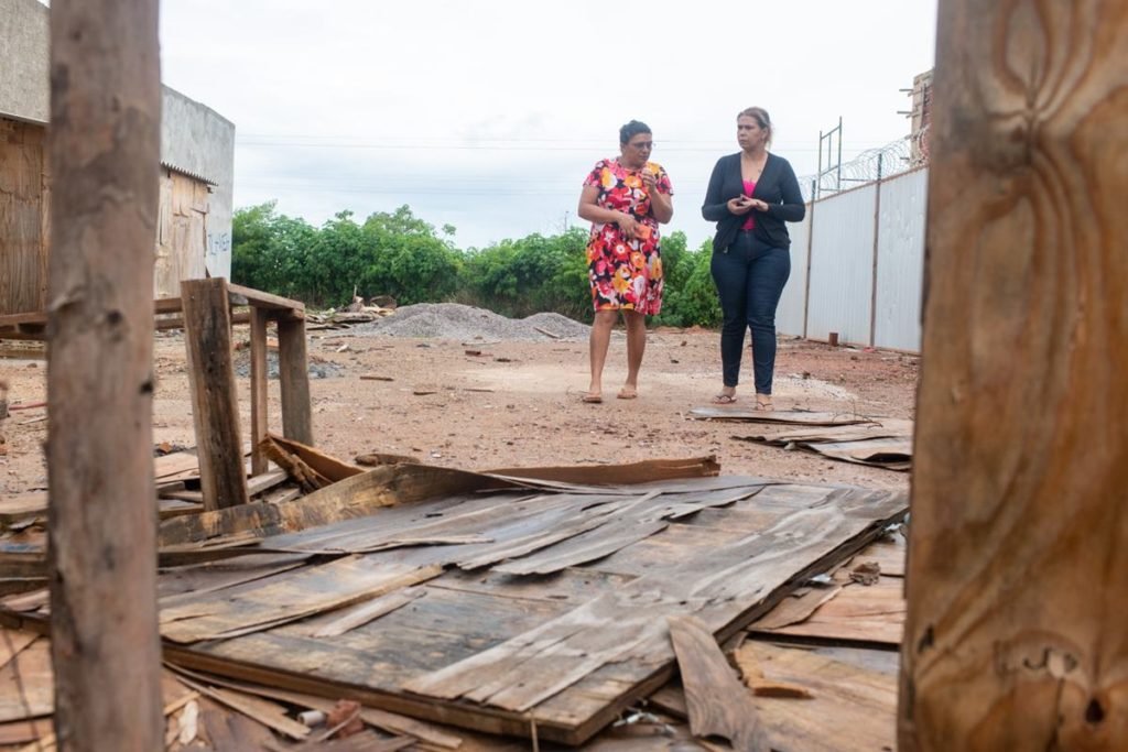 Mulher vestindo casaco preto anda ao lado de outra usando vestido florido. Ambas caminham por um terreno baldio. Foto colorido