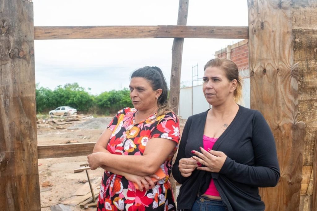 Mulher vestindo casaco preto anda ao lado de outra usando vestido florido. Ambas caminham por um terreno baldio. Foto colorido
