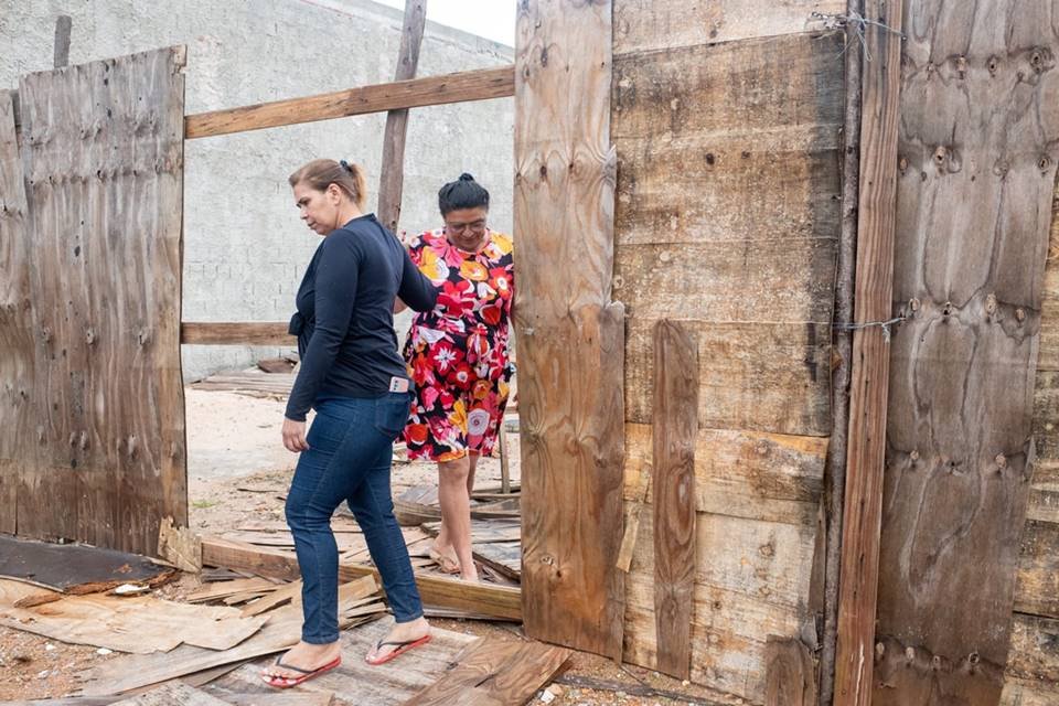 Mulher de costas vestindo casaco preto anda ao lado de outra usando vestido florido. Ambas caminham por um terreno baldio. Foto colorido