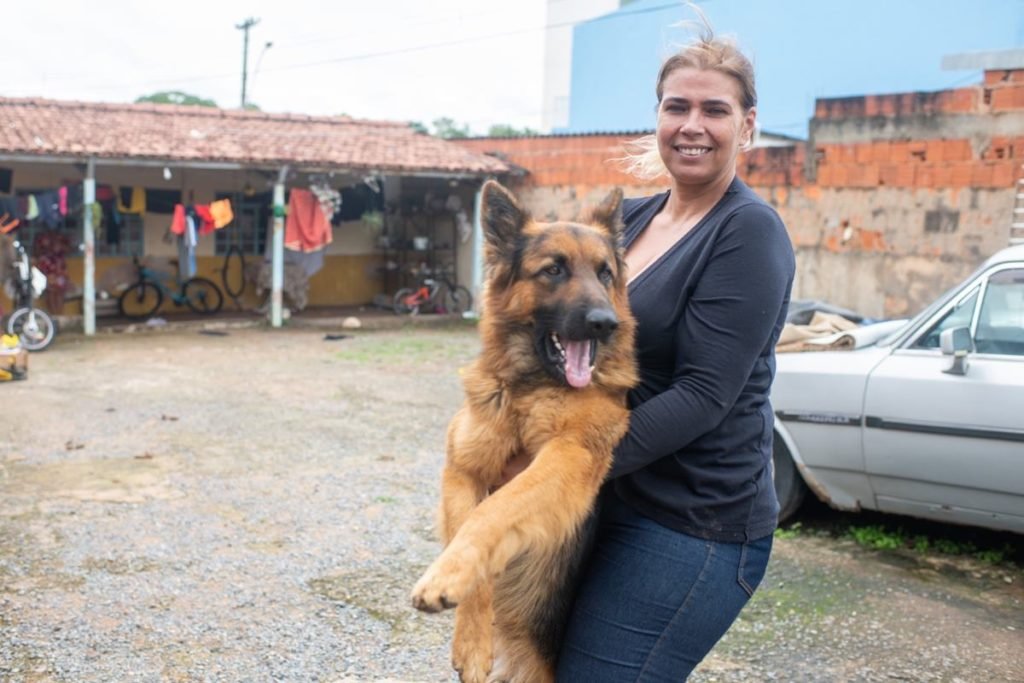 Mulher loira vestindo casaco preto e calça jeans segura pastor alemão. Ao fundo, uma casa de um andar com varal exposto. Foto colorida