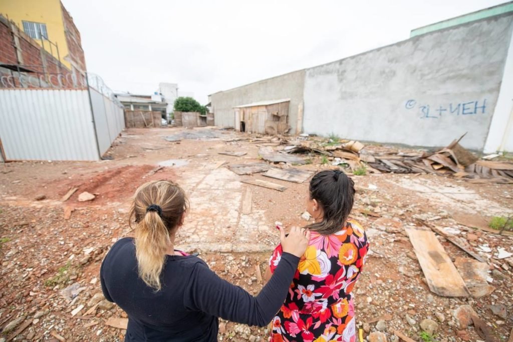 Mulher de costas vestindo casaco preto coloca mão no ombro de outra usando vestido florido. Ambas olham para um terreno baldio. Foto colorido