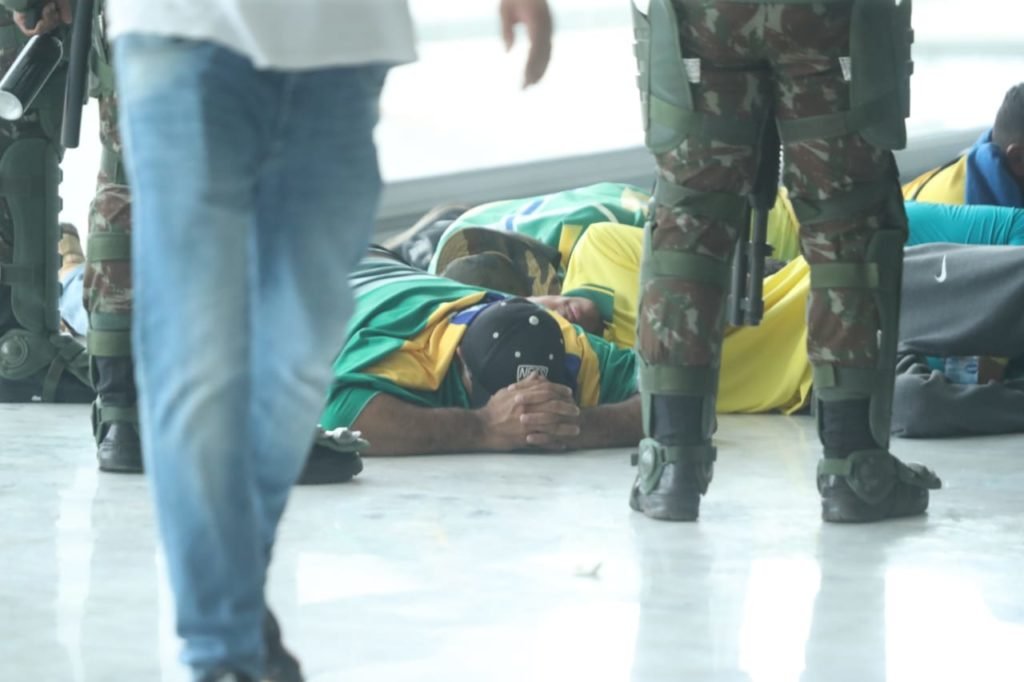 Manifestantes bolsonaristas presos no Palácio do Planalto durante protestos antidemocráticos na Esplanada dos Ministérios. Eles aparecem deitados no chão, enrolados em bandeiras do Brasil enquanto homens fardados caminham ao redor - Metrópoles
