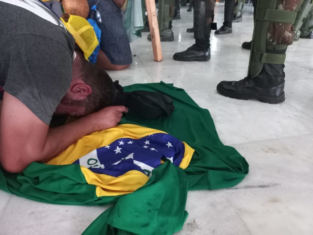 Manifestantes bolsonaristas presos no Palácio do Planalto durante protestos antidemocráticos na Esplanada dos Ministérios. Agentes da força de segurança mantém um homem no chão, que se ajoelha sobre a bandeira do Brasil - Metrópoles