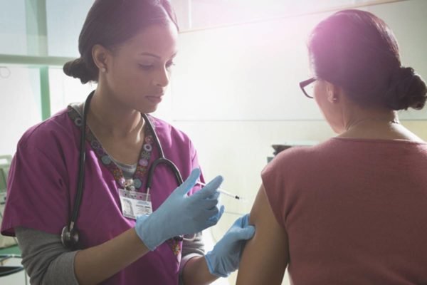 Profissional da saúde aplicando vacina em senhora que está de costas para a câmera