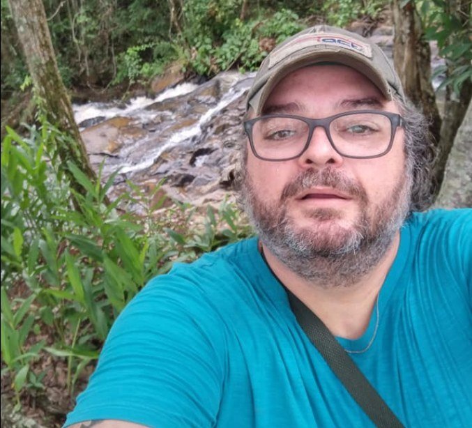 Homem branco, de meia idade, barba grisalha, usa boné e camisa azul. Faz selfie em região de mata. Foto colorida