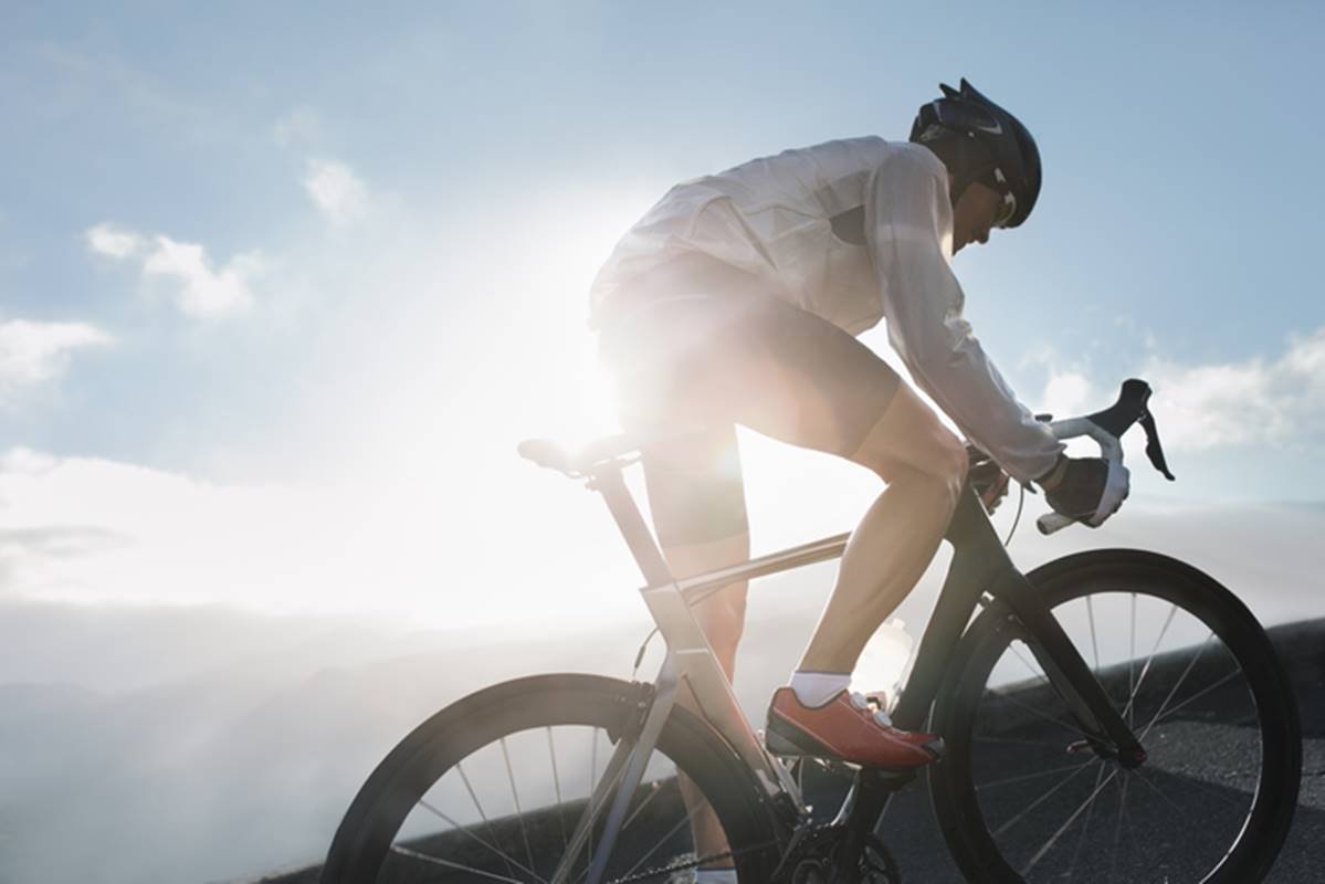 Ciclista pedalando no que aparenta ser uma subida com um sol forte ao fundo da paisagem