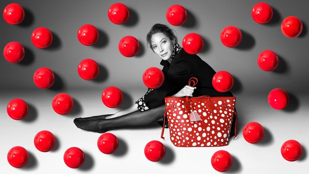 A modelo Christy Turlington, uma mulher branca e de meia idade, com cabelo liso castanho, posa para campanha da marca Louis Vuitton. Ela usa um vestido preto com a gola de bolinhas brancas, meia calça e uma bolsa vermelha com bolas brancas. - Metrópoles