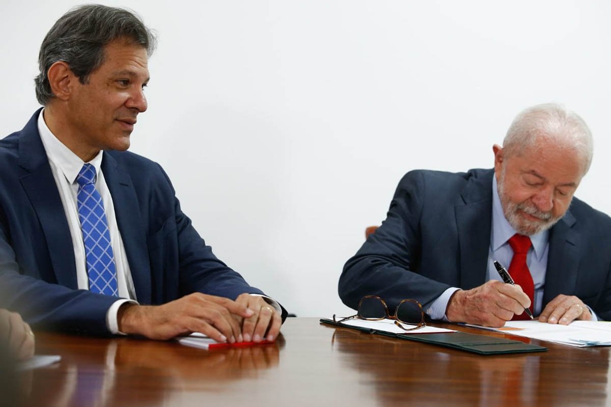 O ministro da Economia, Fernando Haddad, conversa sentado em mesa enquanto o presidente Lula, ao lado, assina documento - Metrópoles