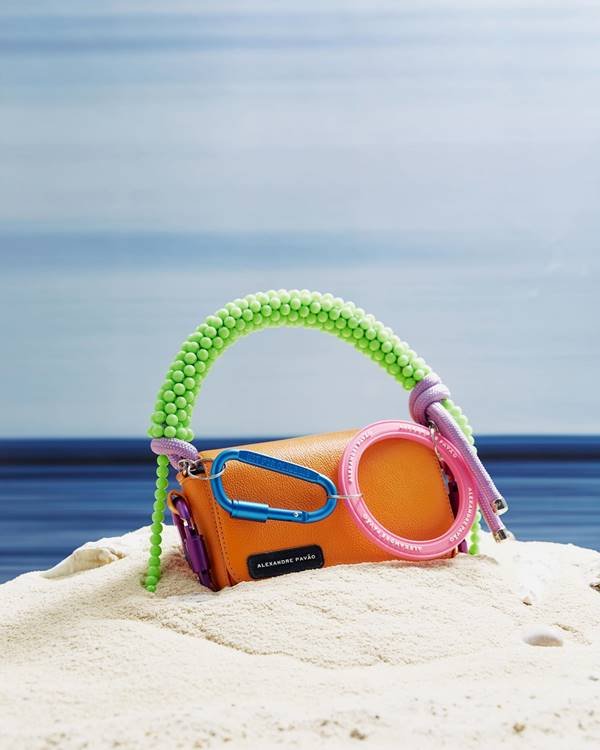 Bolsa da marca Alexandre Pavão nas cores laranja, rosa e verde estão posicionada em cima da areia - Metrópoles