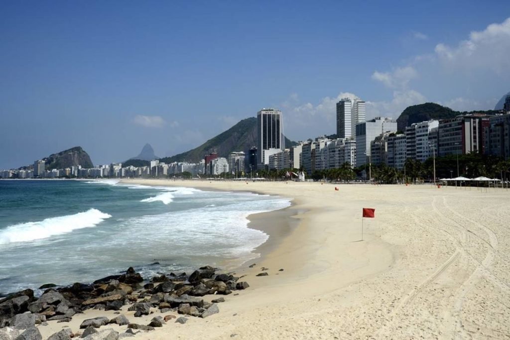 foto colorida de faixa de areia, mar e predios ao fundo - metrópoles