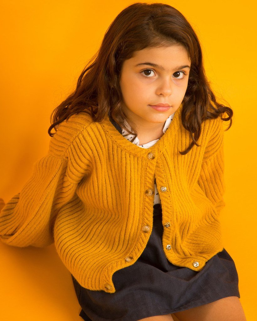 Foto de divulgação da marca de roupas infantis Edamami. Uma menina branca, de no máximo 6 anos, posa para a imagem. Ela possui cabelos castanhos longos e lisos e usa um casaco amarelo de tricô. - Metrópoles