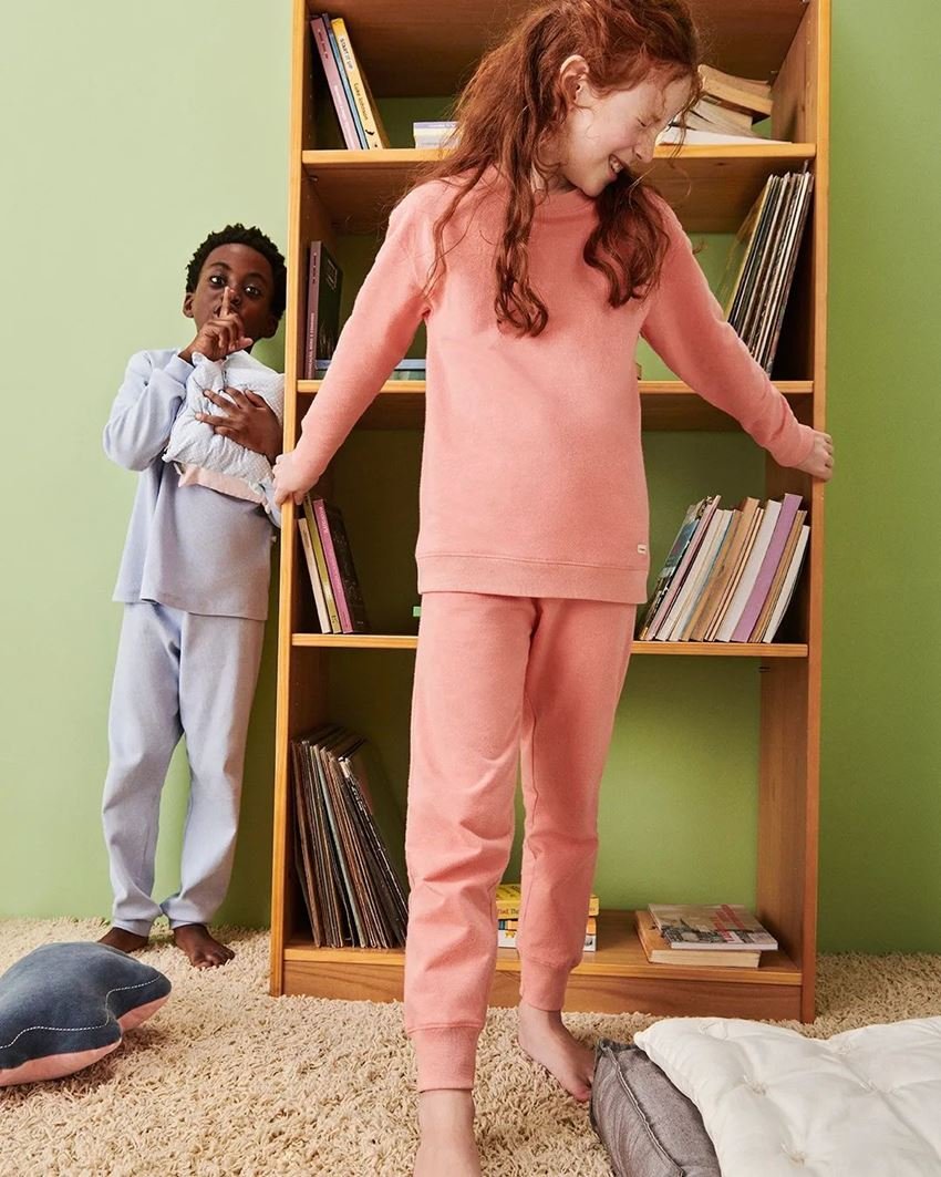 Duas crianças posam para foto de campanha da marca Hering Kids. Um menino negro de cabelo cacheado usa um conjunto de casaco e calça, ambos de moletom, na cor cinza. A menina branca, de cabelos lisos ruivos, usa um conjunto de moletom rosa. - Metrópoles
