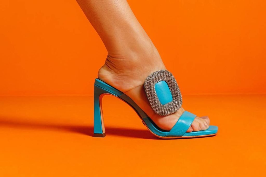 Foto com foco no pé da modelo. A modelo usa uma sandália com detalhe brilhoso em um fundo laranja - Metrópoles