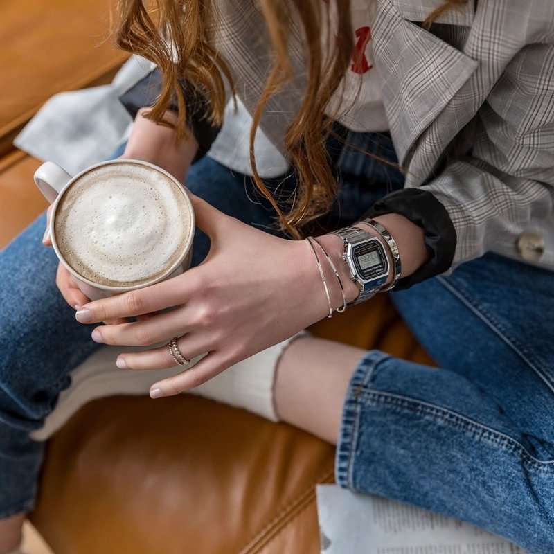 Mulher branca e jovem, de cabelo liso castanho, posa para foto.  Ela usa um casaco cinza, calça jeans, meias brancas e um relógio dourado da marca Casio.  - Metrópoles